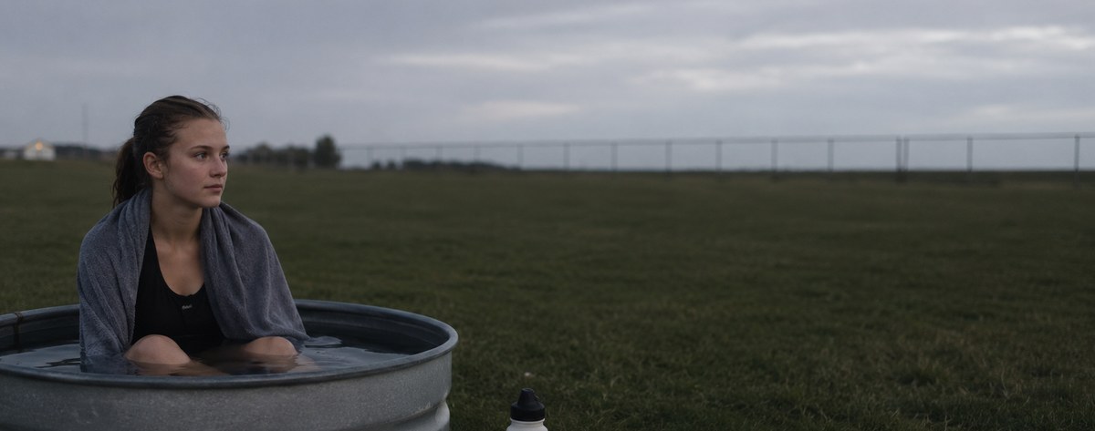 A high-school-age distance runner sitting in a plain galvanized stock tank of cold water on a practice field at dusk, towel over her shoulders, calm and unremarkable — no ice, no fog, no dramatic lighting.
