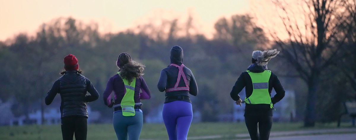 Four runners seen from behind, jogging easy on an open road at dawn, relaxed and unhurried, with the sky glowing on the horizon.