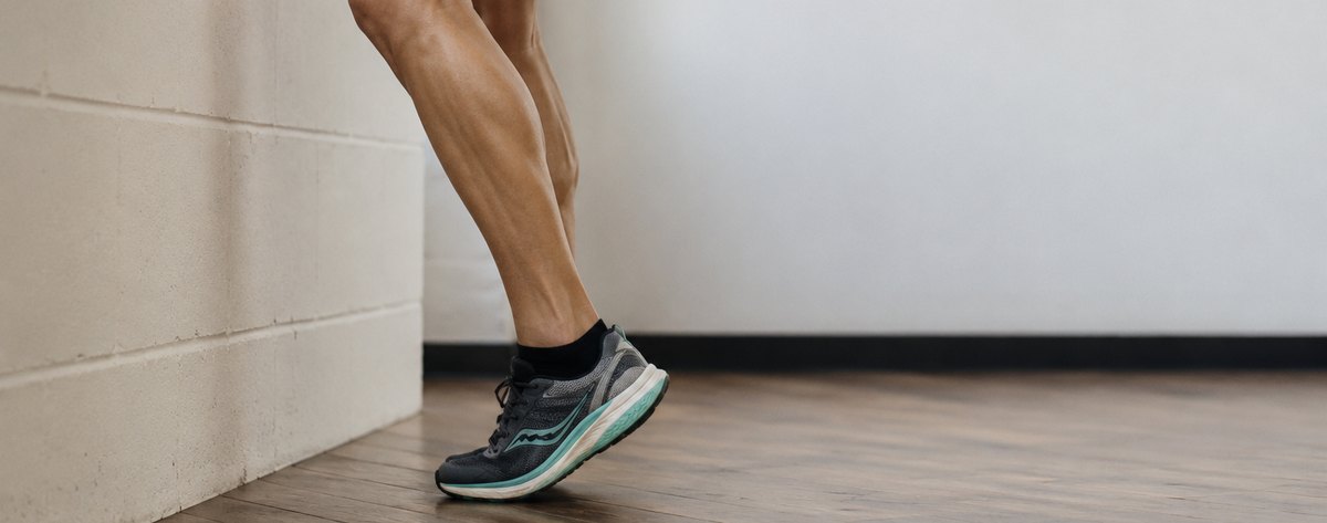 A runner performing a single-leg isometric calf raise hold against a wall, viewed from the side in a gym.