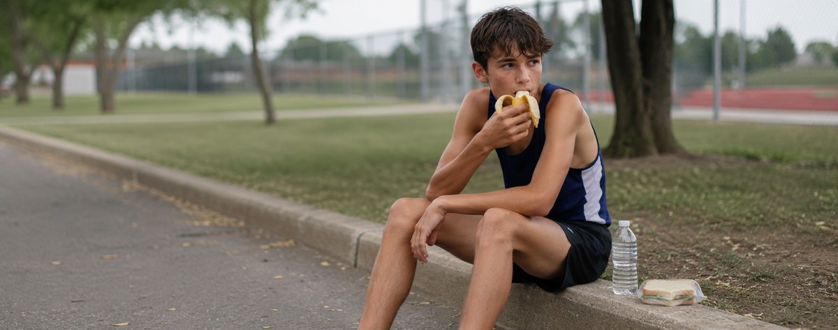 A high-school cross-country runner sitting on a curb after practice, eating a banana and a sandwich out of a backpack.