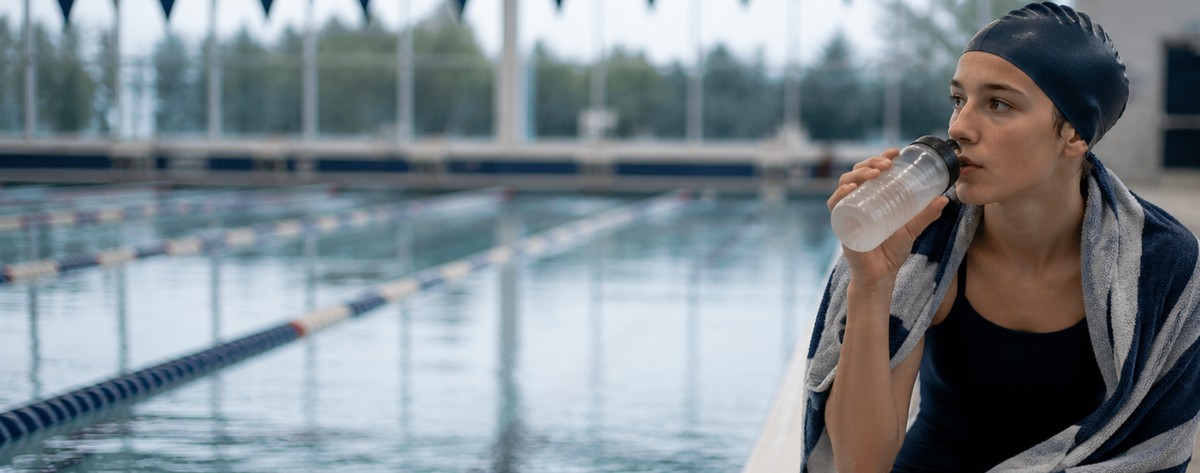 A high-school swimmer sitting on the pool deck at dawn wrapped in a towel, eating a banana and drinking from a water bottle before an early-morning practice.